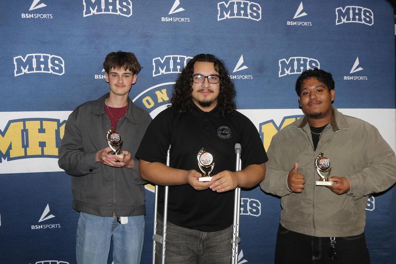 Three high school students stand in front of a blue MHS and BSN Sports backdrop, each holding a small trophy. The student in the center, who is using crutches, wears a black shirt, while the others wear light jackets. All three smile proudly, with the student on the right giving a thumbs-up.