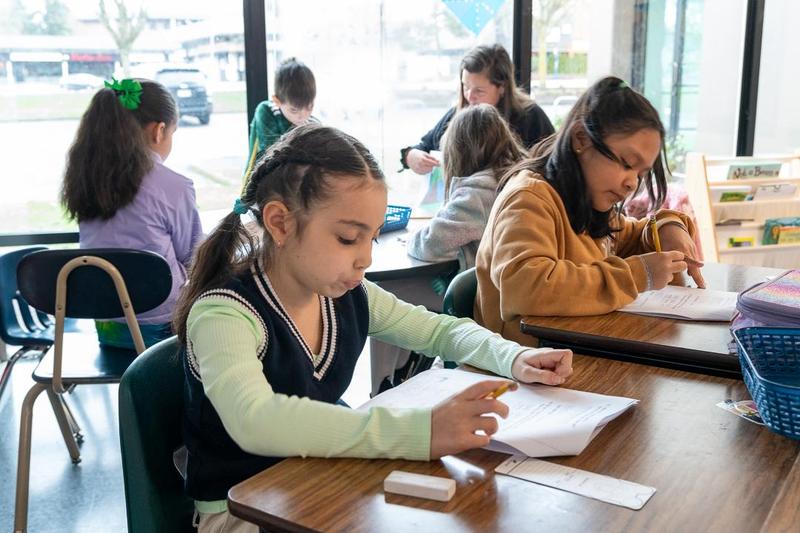 Arco Iris students and an adult sit at desks in front of full-length wall windows.