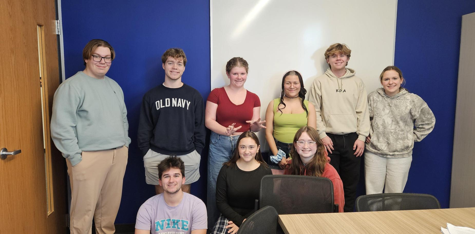 A group of nine young people posing together in a classroom setting.