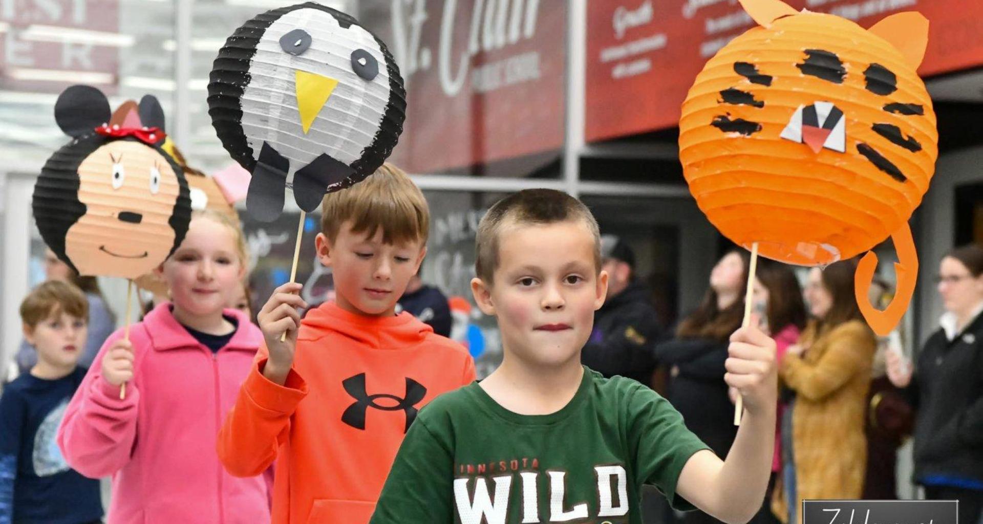 Children holding decorated lanterns in a festive parade inside a building.