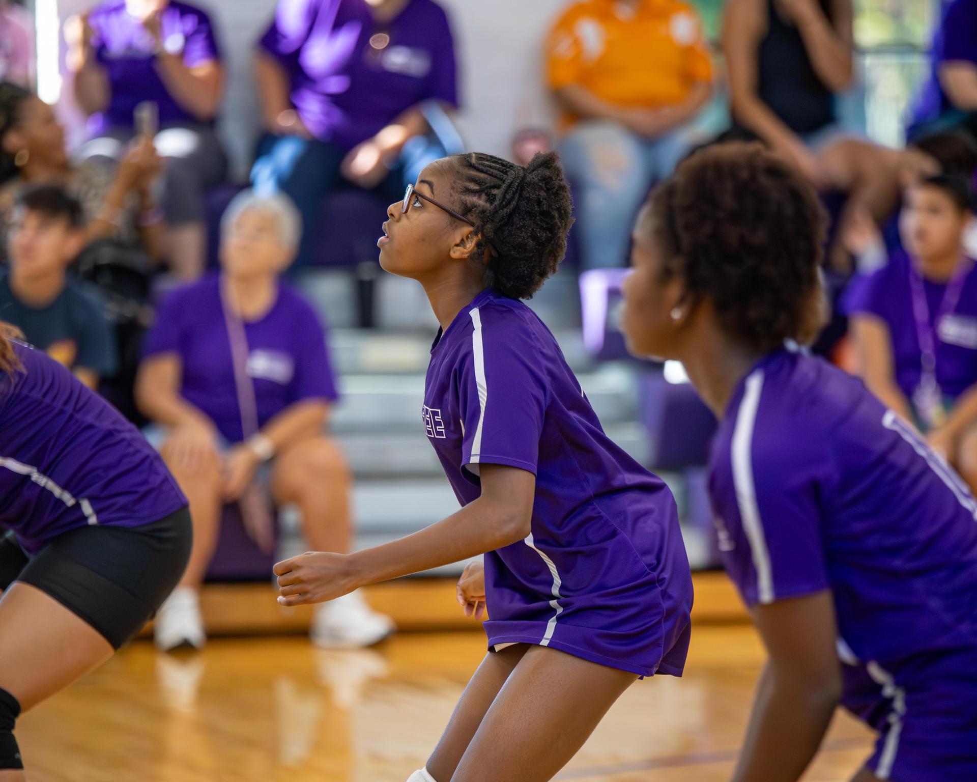 Young players in purple shirts prepare for a volleyball play in a gym, focused and attentive.