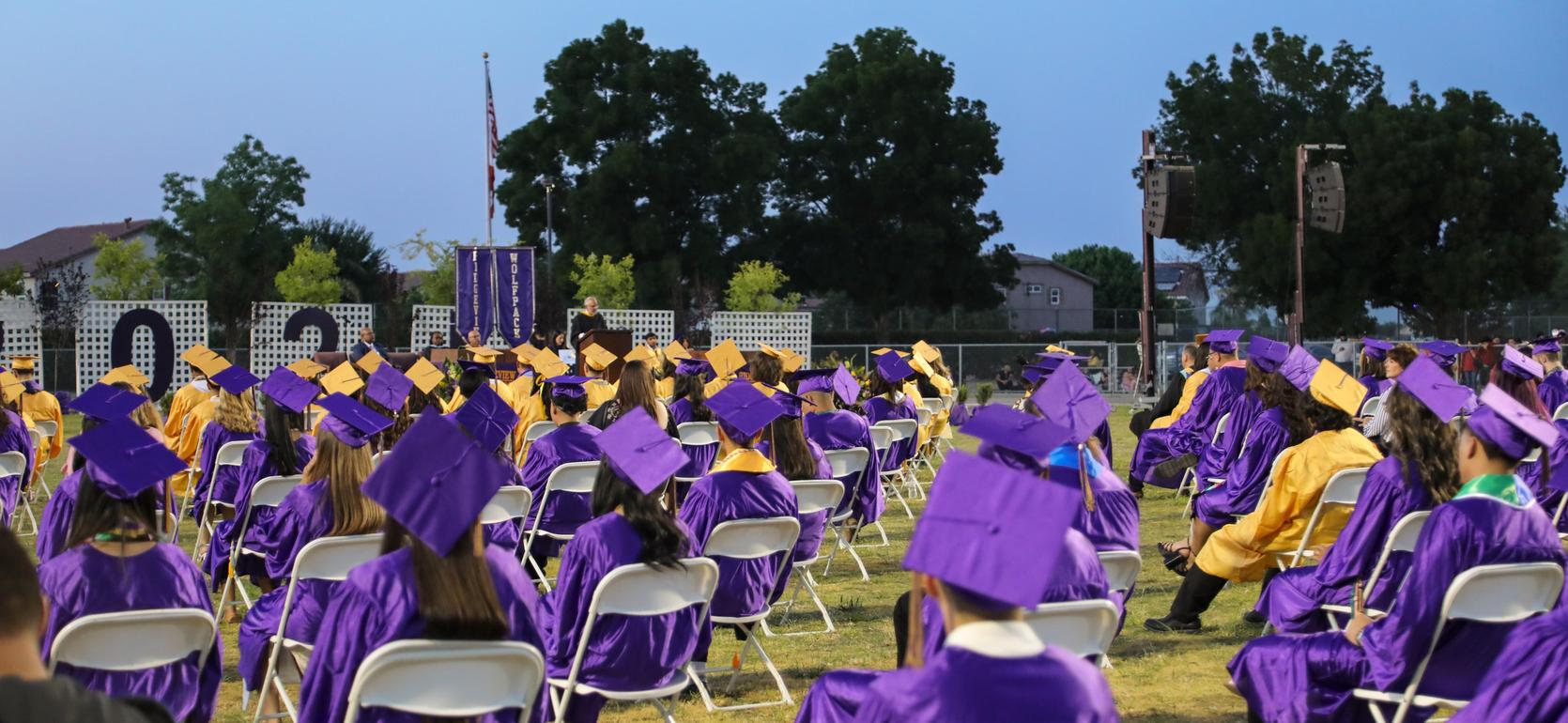 Gathering of graduates in purple and gold gowns, seated in rows, with audience visible behind.