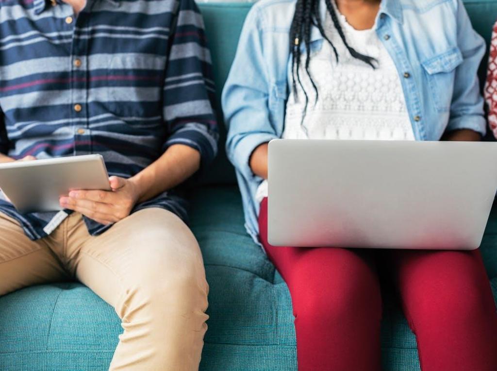 Two individuals sitting on a couch, one with a tablet and the other with a laptop.