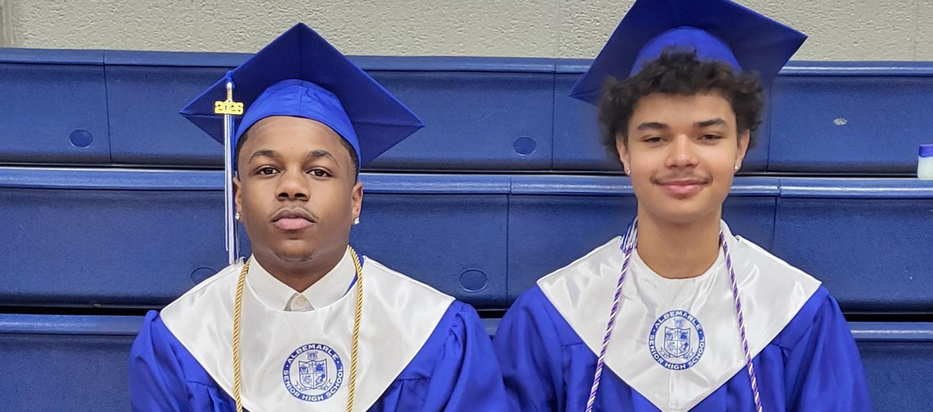 Two graduates in blue gowns sit closely together, displaying their attire and accessories.