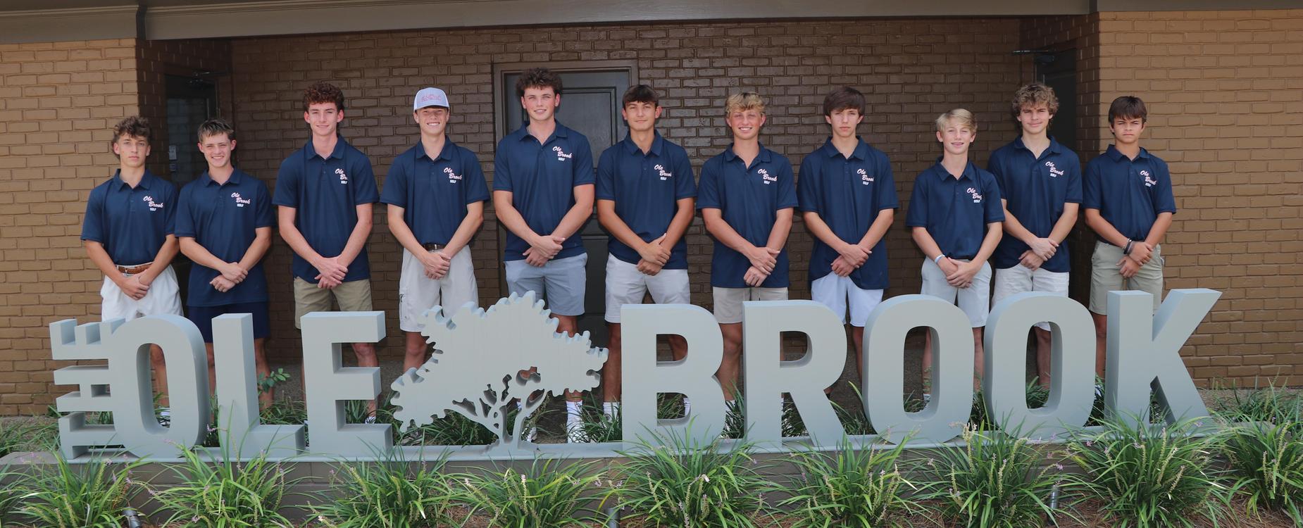 A group of eleven young men in matching polo shirts standing by a large sign that reads 'THE OLE BROOK' with greenery around.