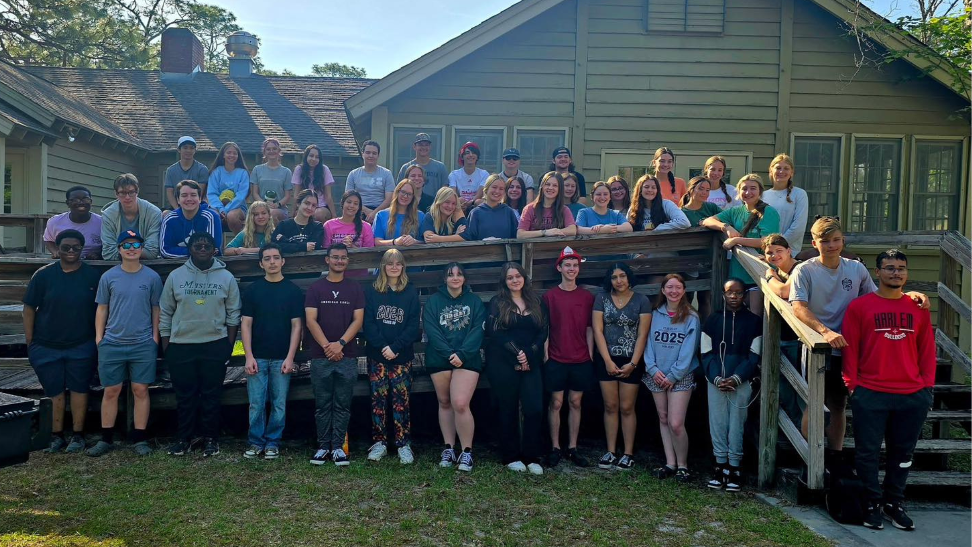 A large group of students smiling on a deck in front of a wooden building.