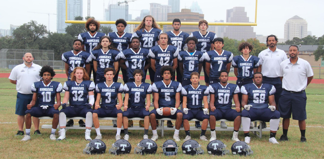 Team photo of the Texas School for the Deaf (TSD) 2020 Football State Champions. The group features players in navy blue uniforms with 'RANGERS' written across the chest, standing and sitting on bleachers on a grassy field with a football goalpost and a city skyline in the background. Coaches in white polo shirts and navy shorts stand alongside the team, with helmets placed in front of the seated players.