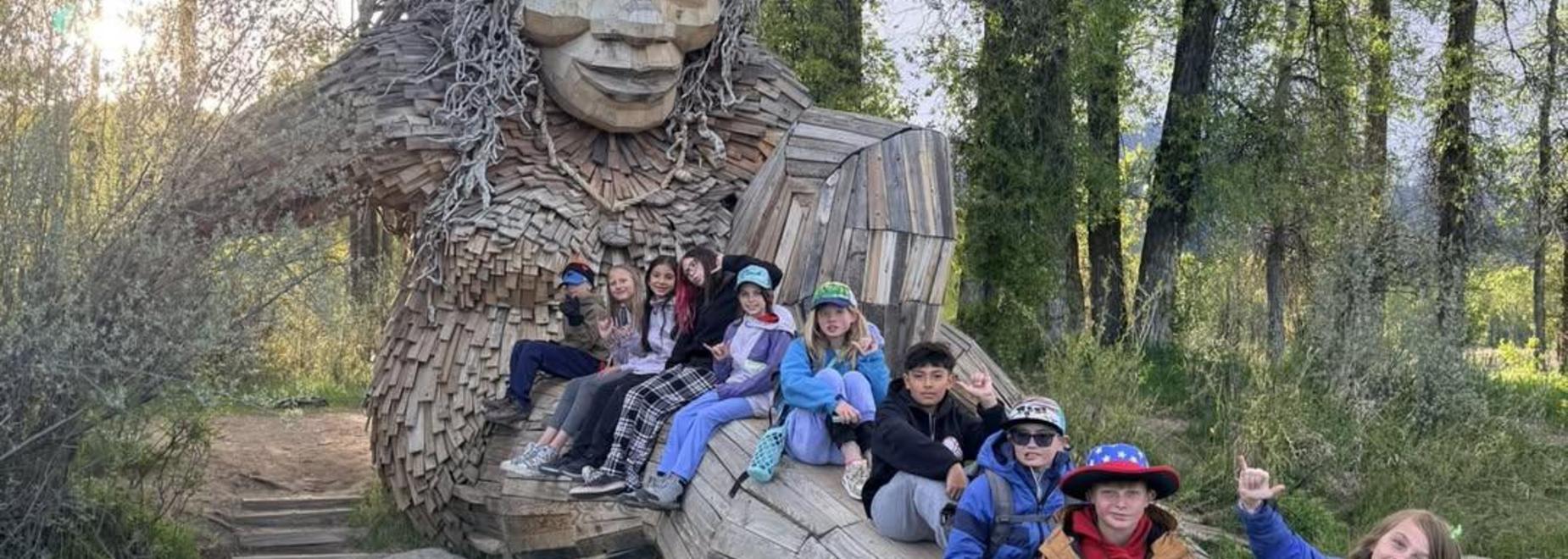Group of children posing by a large wooden sculpture in nature.