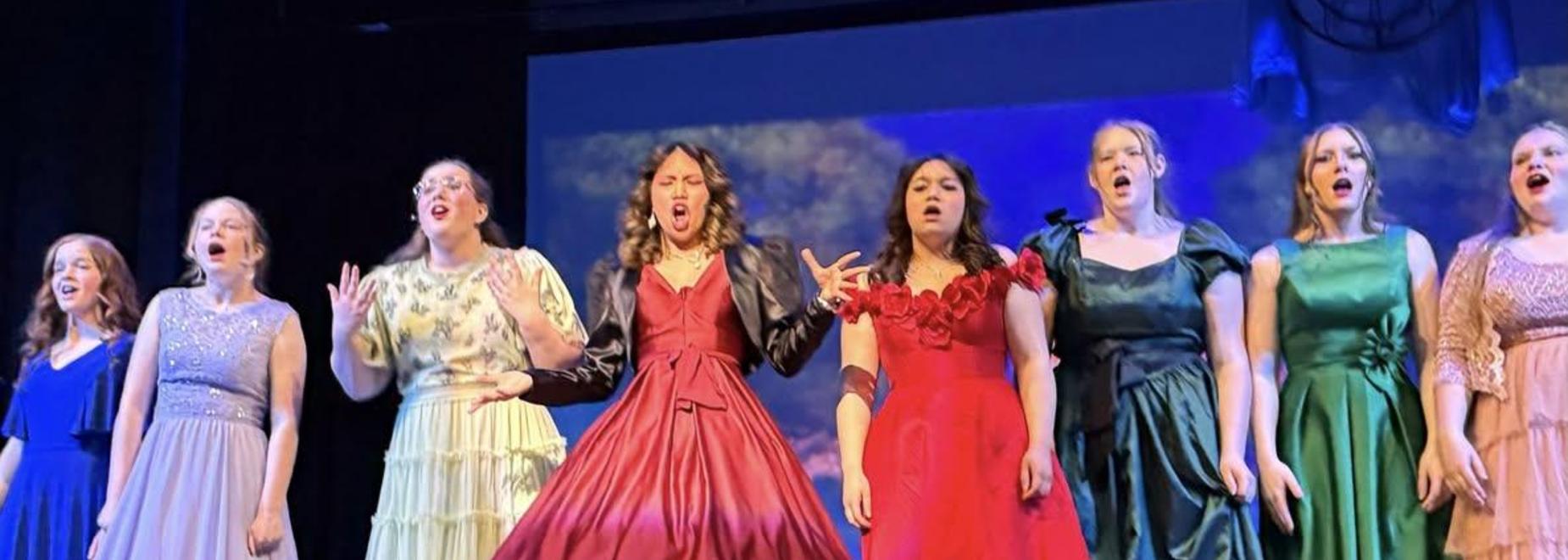 A group of women in colorful gowns singing passionately on stage.
