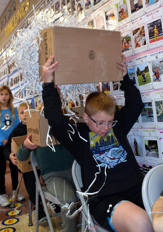 A child lifting a box filled with shredded paper while others watch in the background.