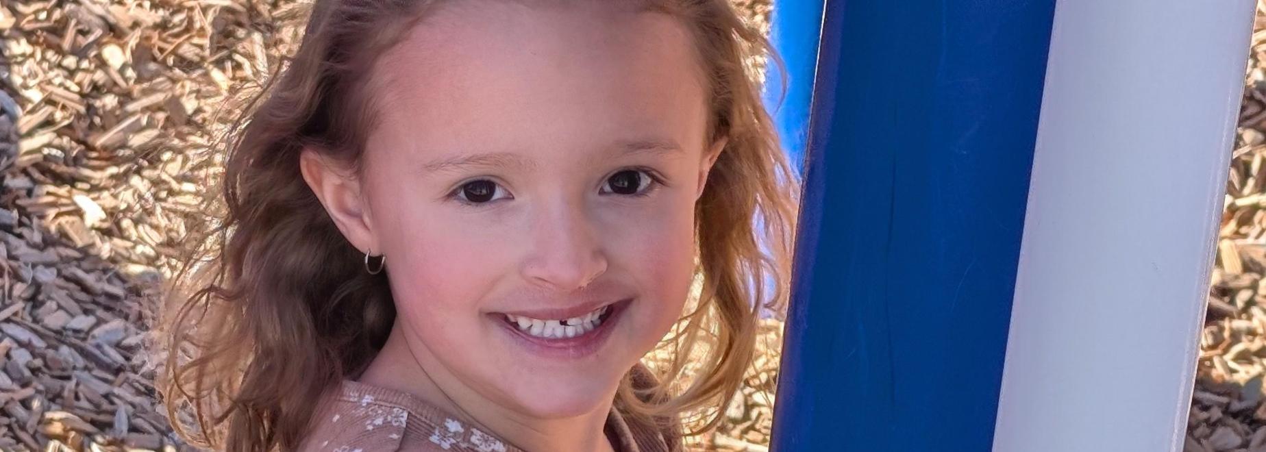 Young girl in a floral shirt holding onto a blue post at a playground.