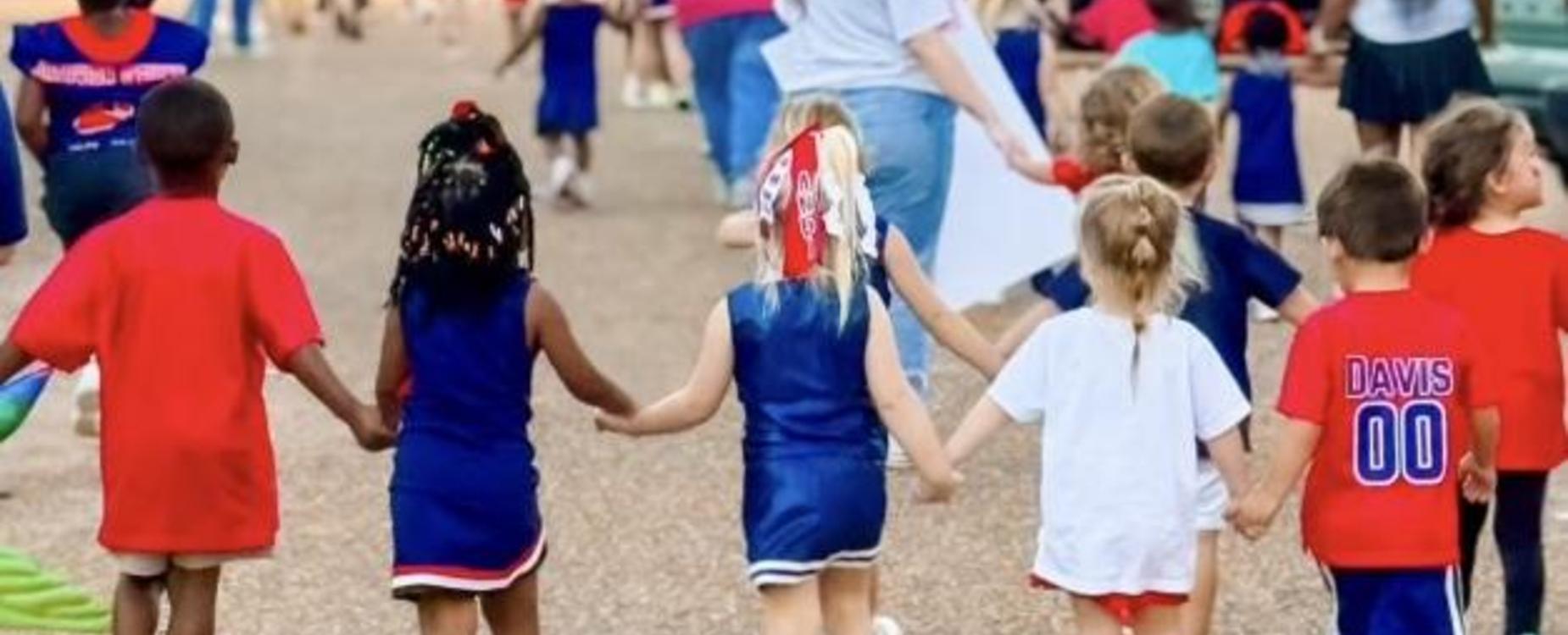 Children holding hands in colorful outfits walking together at an event.