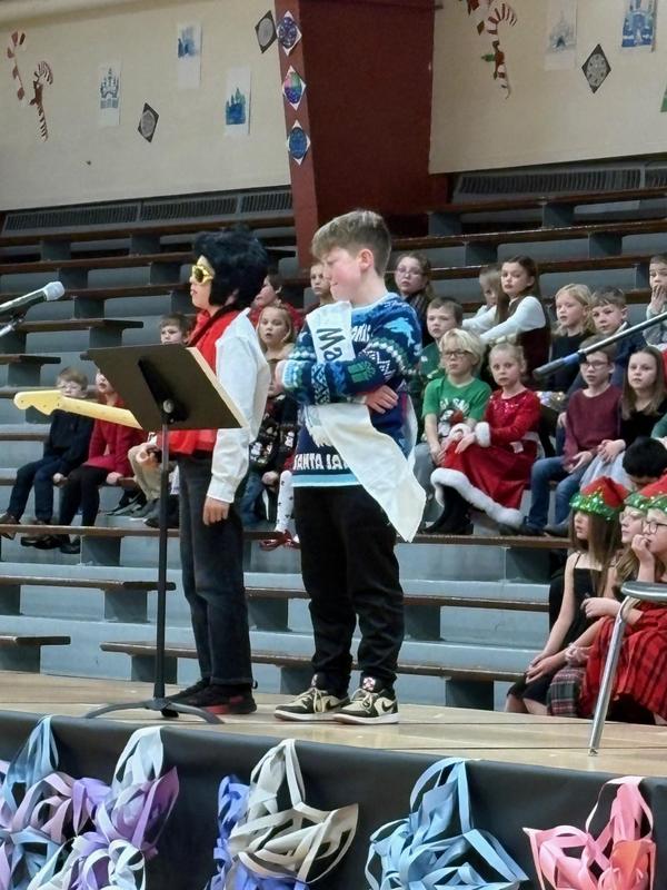 Two performers in costumes on stage during a school event with students watching.