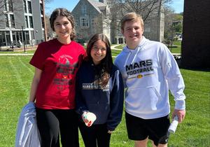 Three students smiling outdoors, one holding a drink, with buildings in the background.