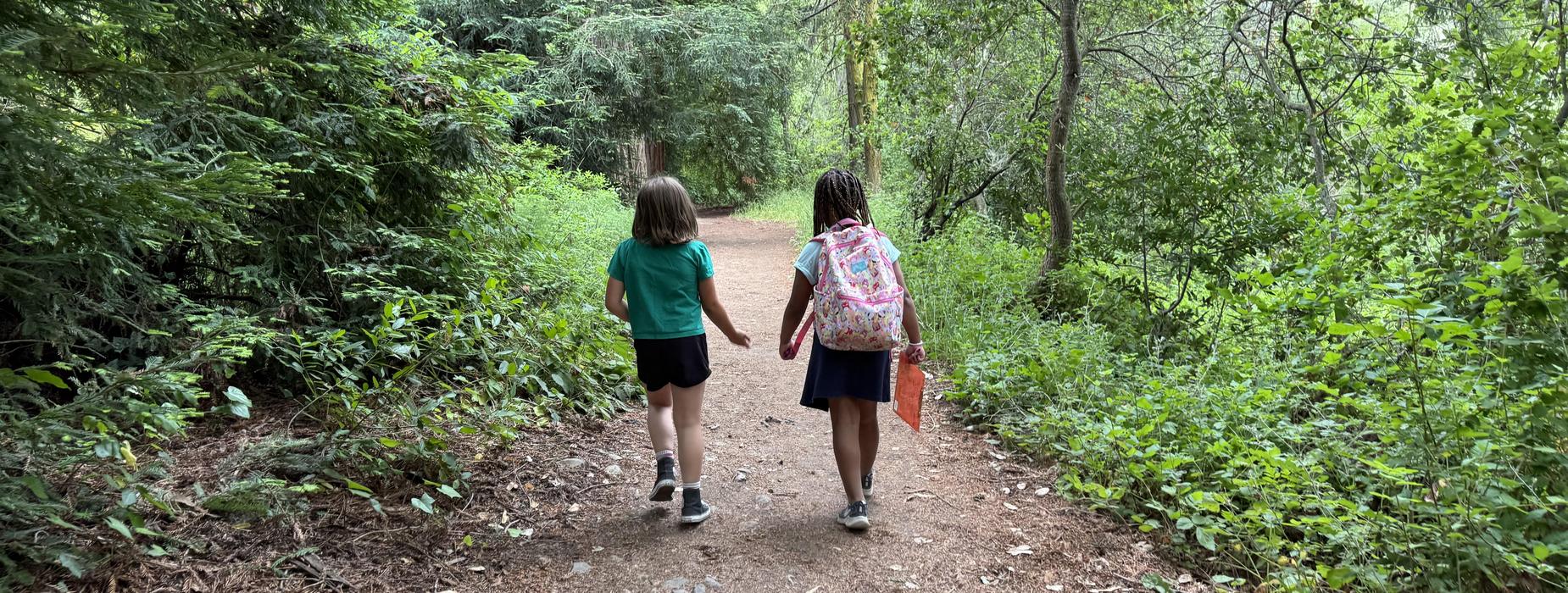 Two children walking together on a dirt path through green trees.