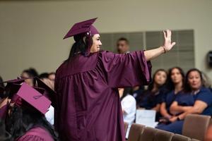 Graduate in a maroon cap and gown smiles and waves to the audience while standing among seated classmates during the Porterville Adult School ceremony.