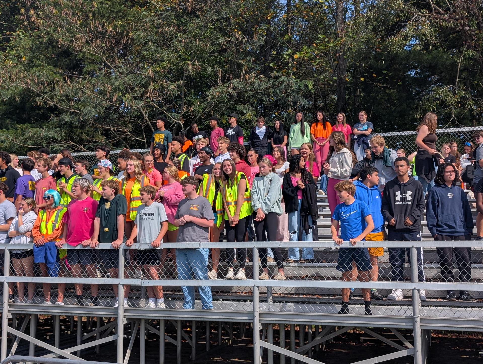 A large group of students in colorful attire watching an event from bleachers at the stadium.