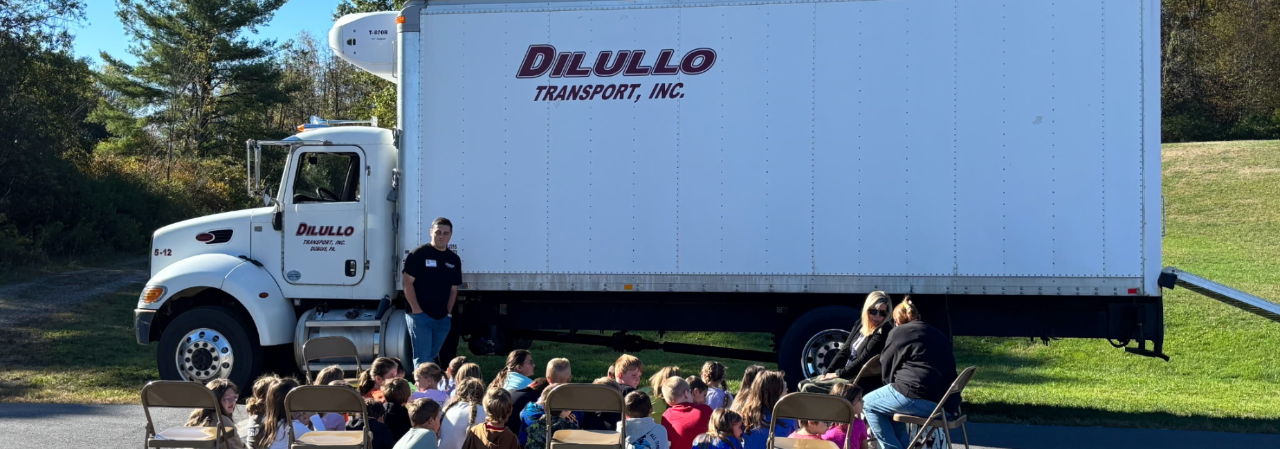 A truck displaying DiLullo Transport, hosting a group of children seated for a talk.