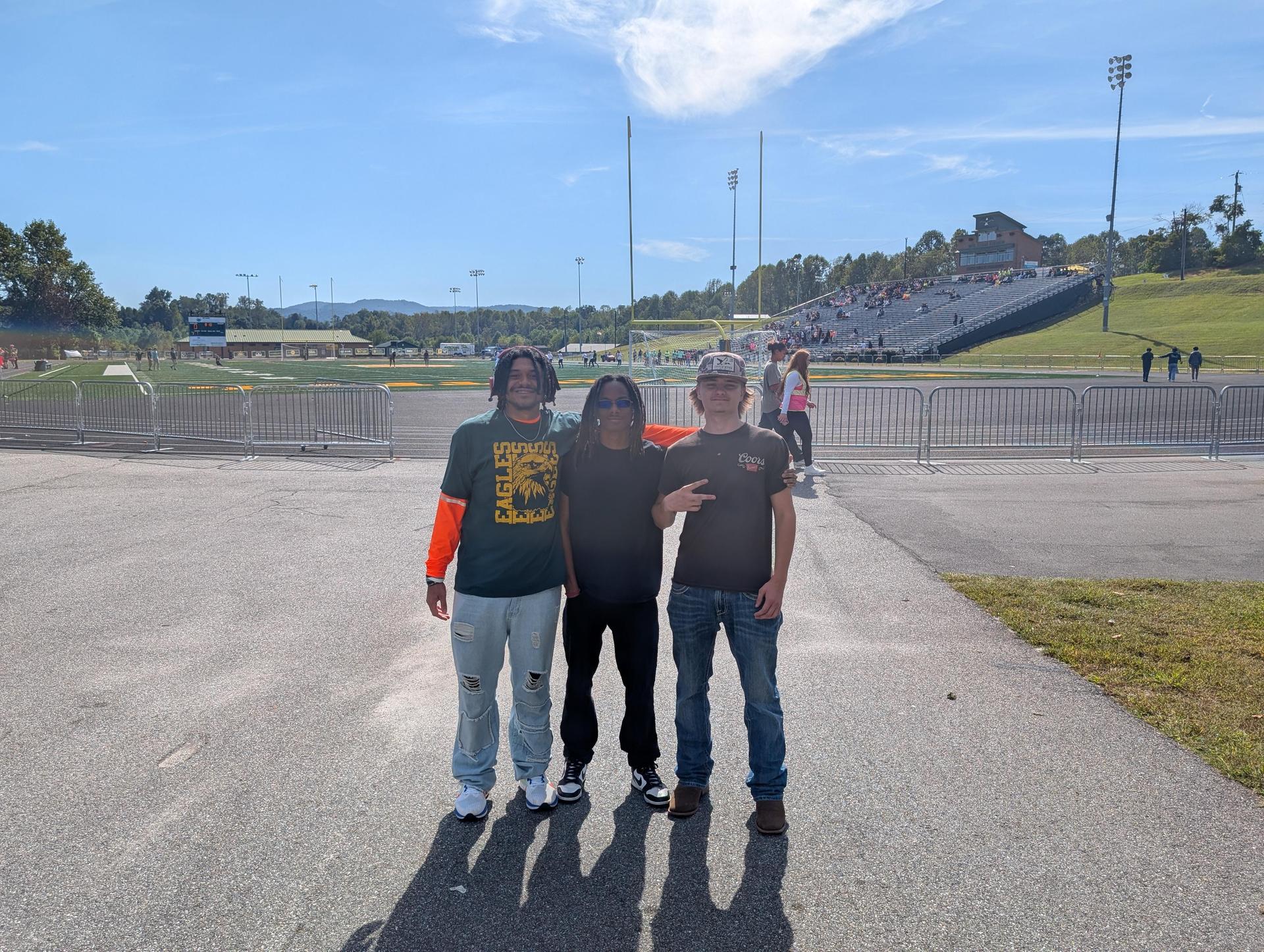 Three friends posing together at an outdoor event with a stadium in the background.