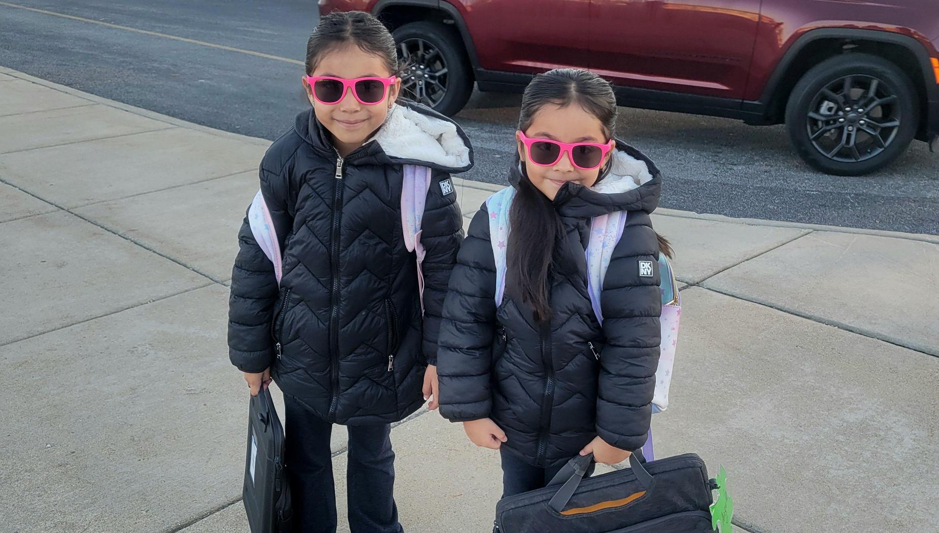 Two girls wearing matching black jackets and pink sunglasses, standing with backpacks.
