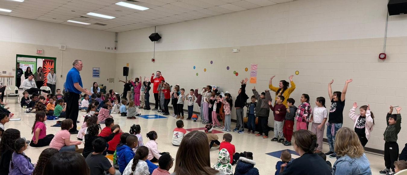 A large group of elementary students raise their hands during an energetic school assembly.