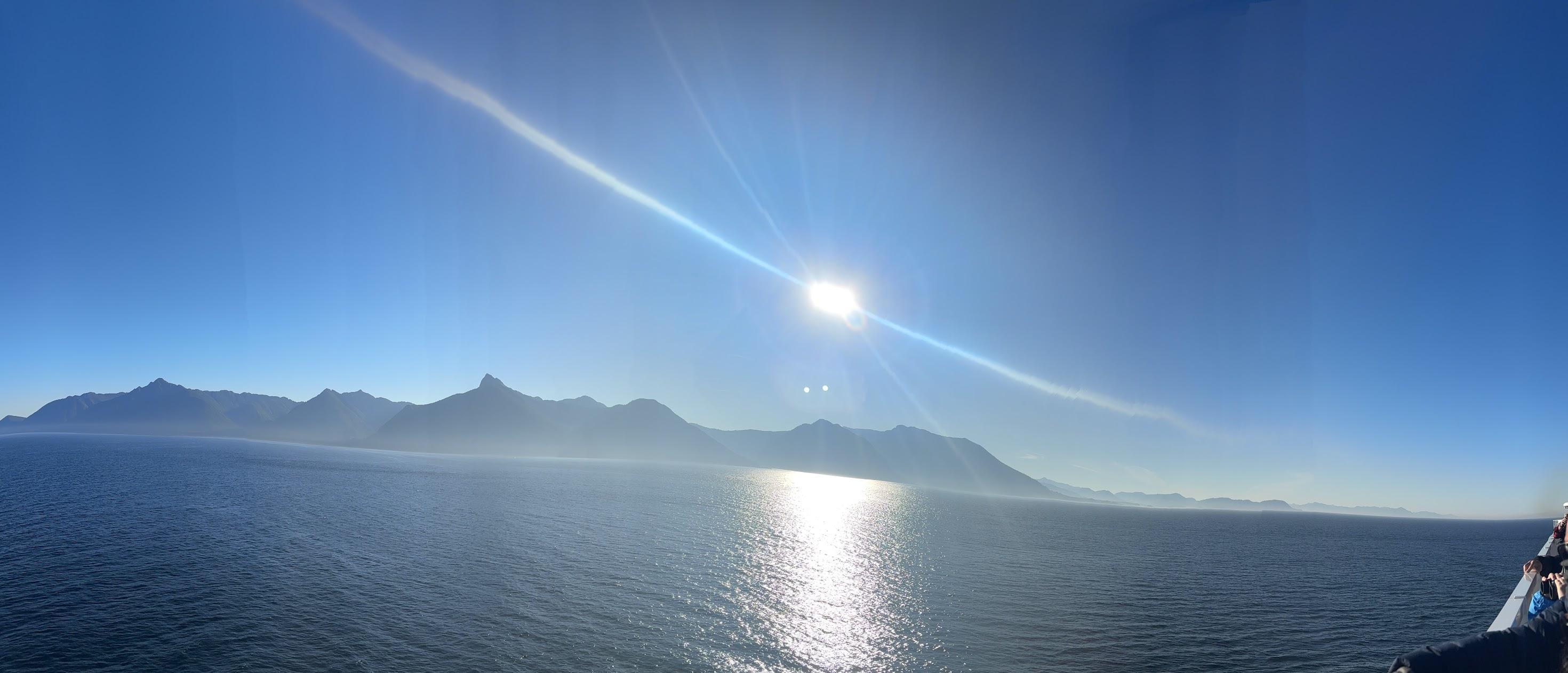 Picture of the sunrise in Southeastern Alaska, 2025. The sun is rising over a clear blue sky. There is water in the foreground, and some mountains in the background. 