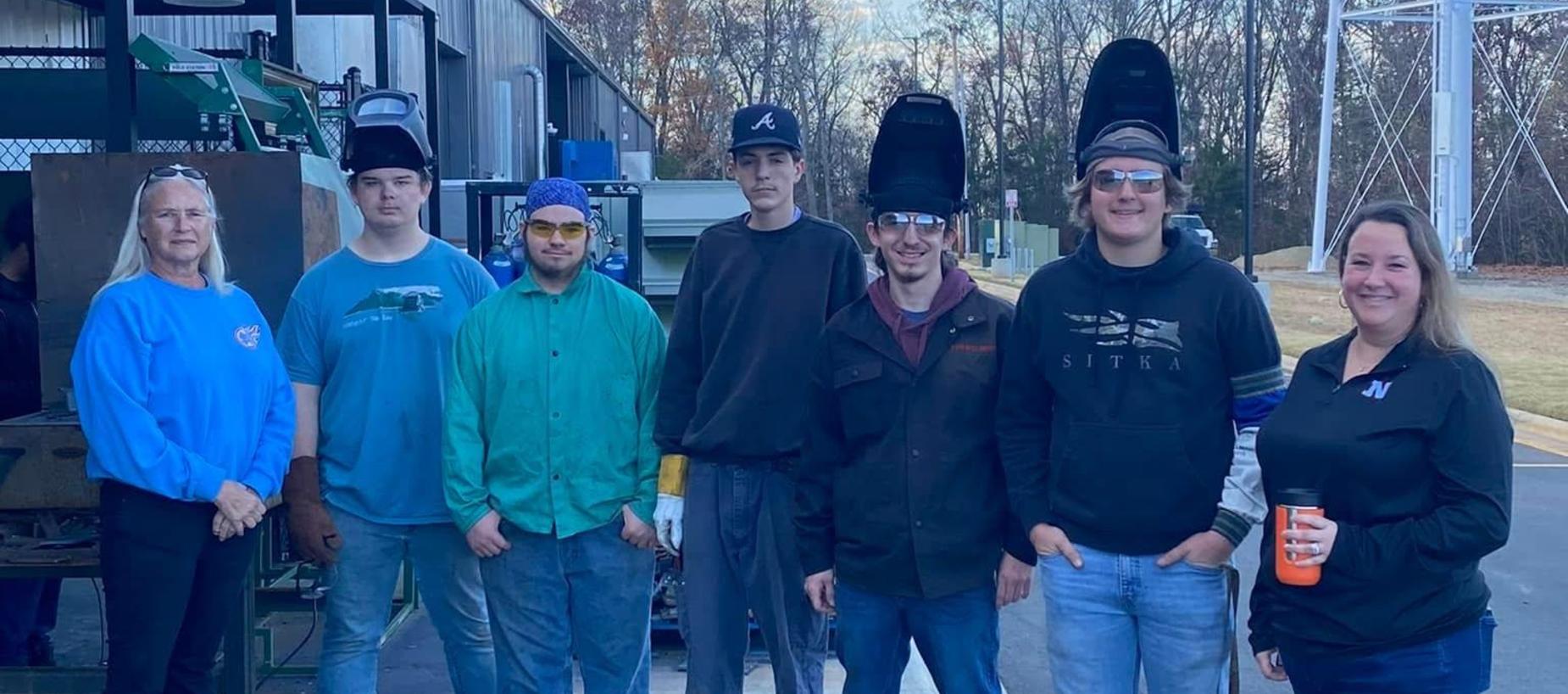 Seven individuals posing in welding gear outdoors, with equipment in the background.