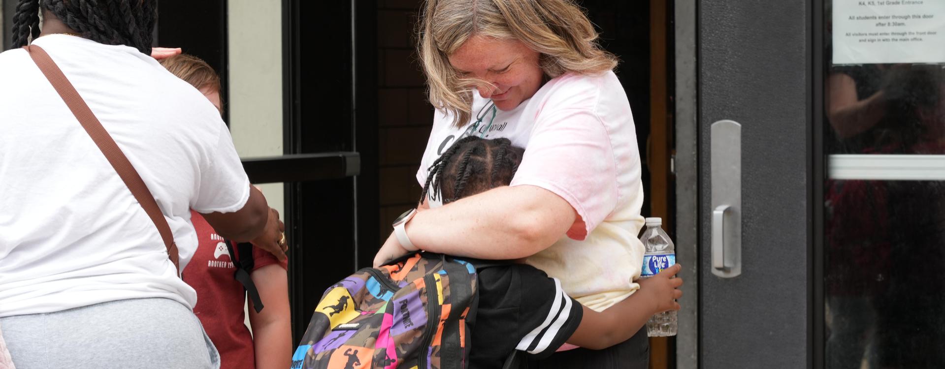 A child hugs an adult while holding a water bottle, outside a building.