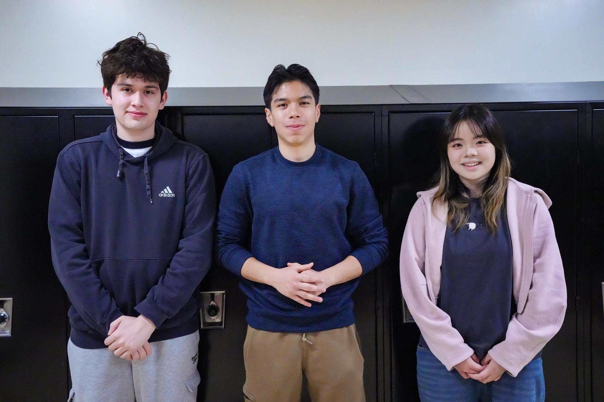 Three students stand together in front of lockers, smiling at the camera.