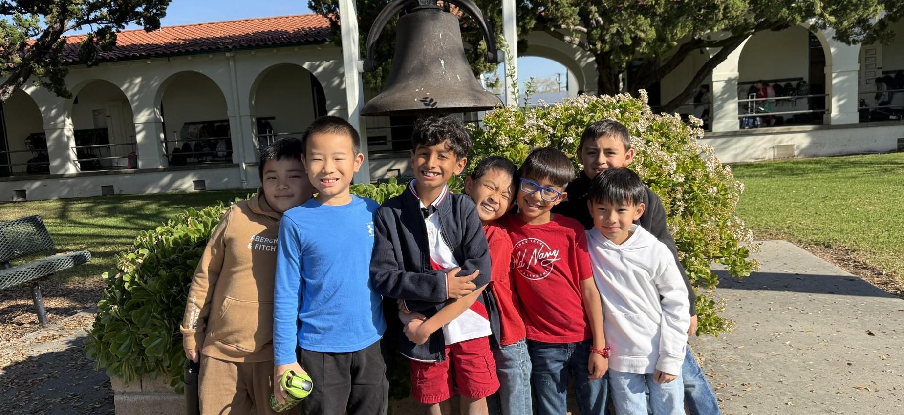 A group of eight boys posing playfully near a bell on a school campus.
