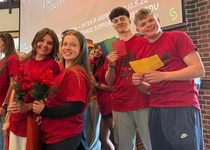 Four smiling students in red shirts hold flowers and colored paper indoors.