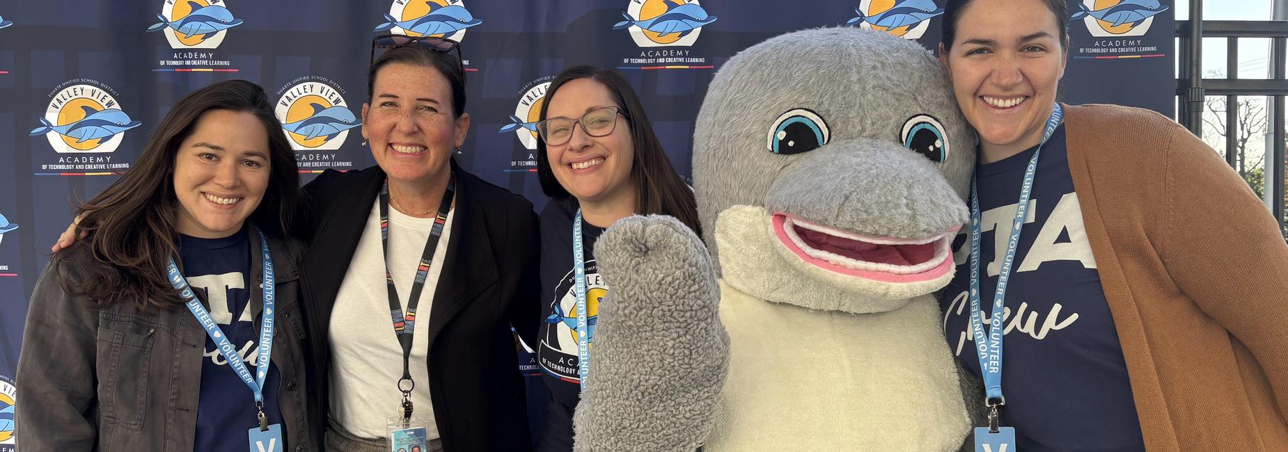 Four women smiling with a dolphin mascot at an event.