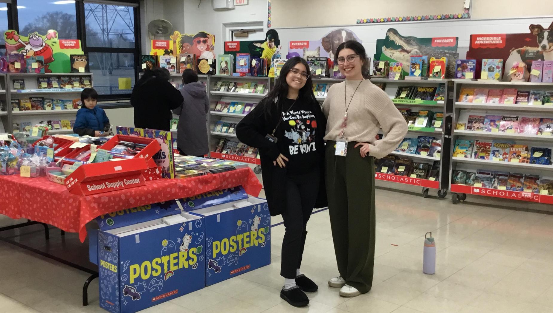 Two smiling women stand in a school supply center surrounded by educational materials.