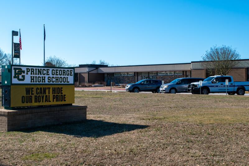 The front of Prince George High School's campus is shown, with vehicles parked at the building near its marquee.