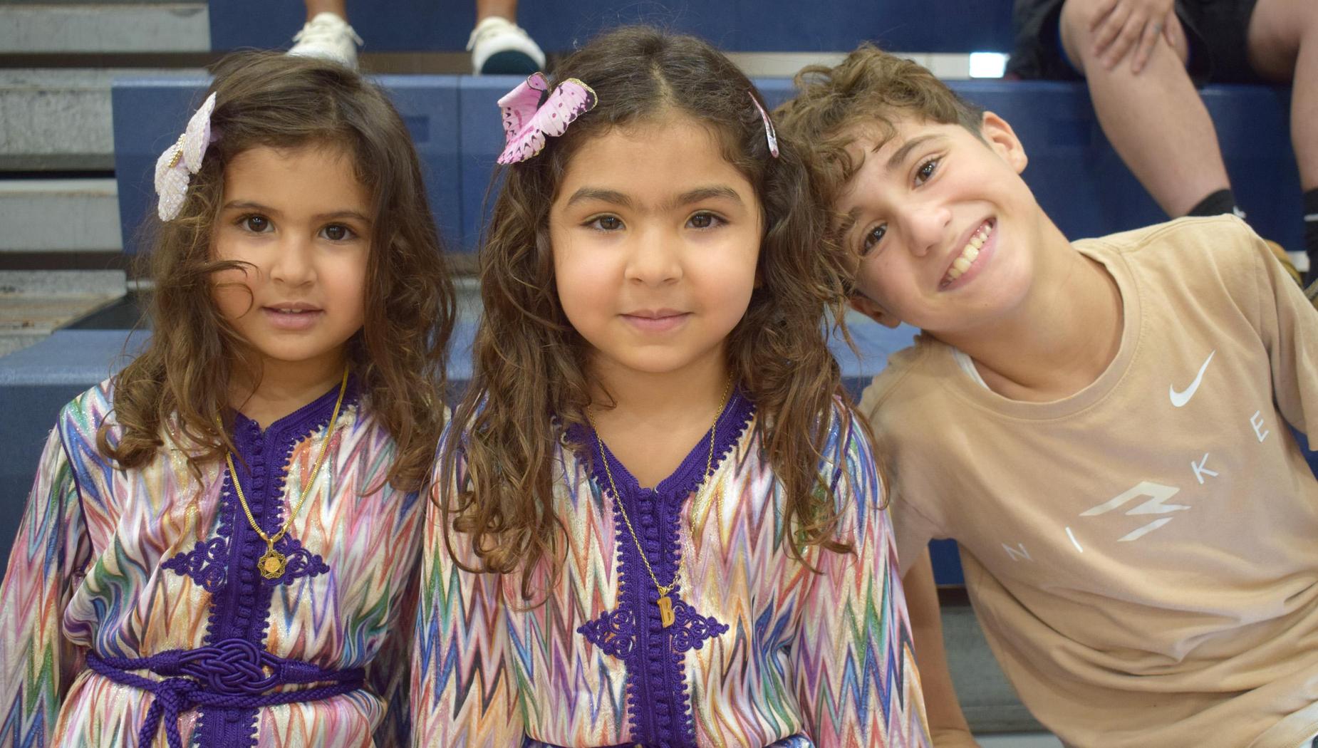 Two girls in colorful dresses and a boy smiling, seated on bleachers.