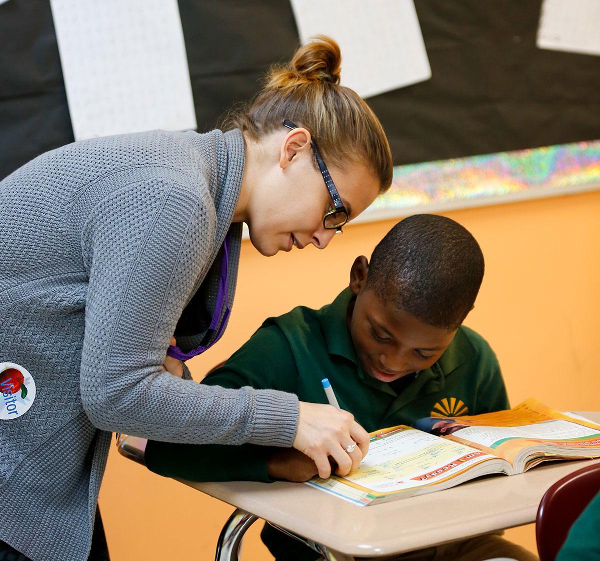 HSA Teacher smiles while kneeling beside a young student in a classroom setting.