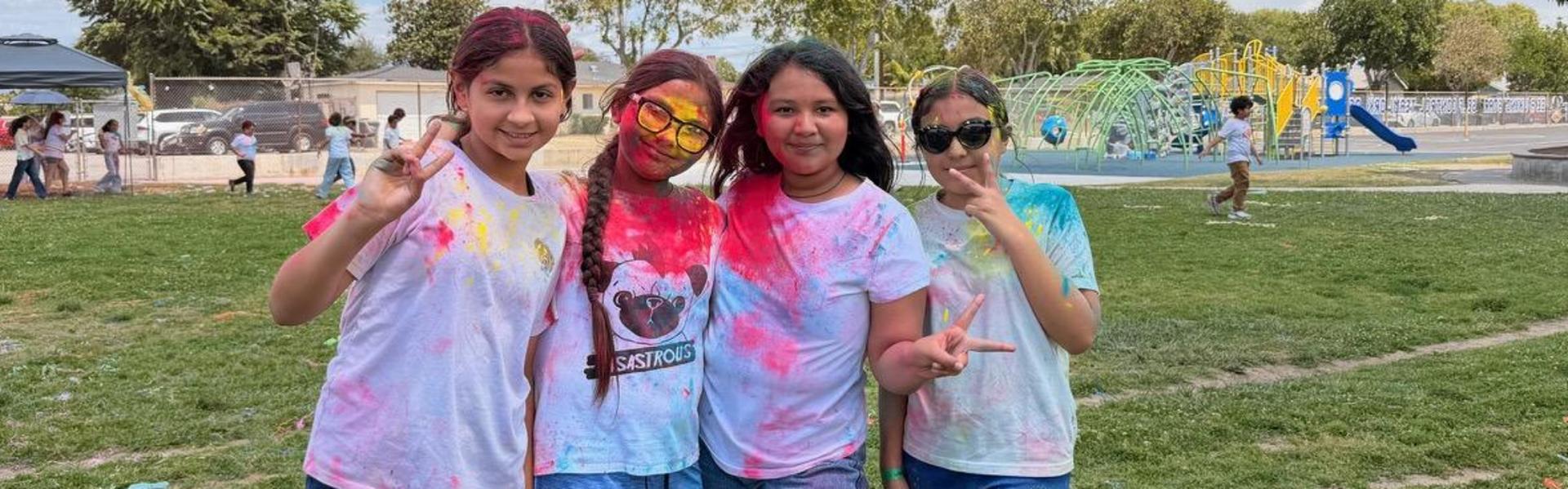 Four girls covered in colored powder, making peace signs in a green park.