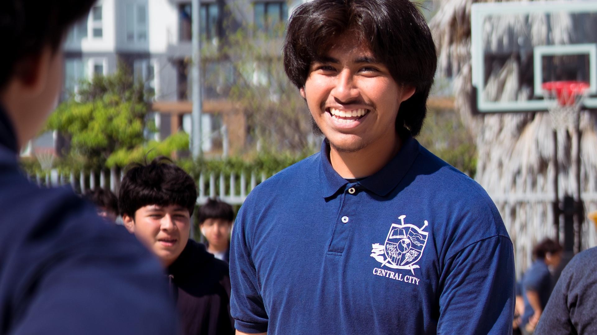 A smiling student in a blue polo shirt greeting others outdoors, with classmates in the background.