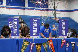 Four students signing commitment letters with confetti and banners in the background.