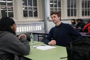 Two individuals having a conversation at a table with a drink and papers.