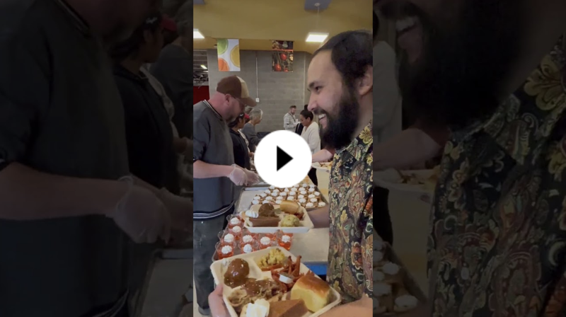 Adult smiling as he receives a full Thanksgiving-style meal from staff serving trays of food in a busy cafeteria.