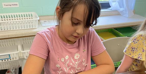 A girl in a pink shirt drawing with a pencil at a table.