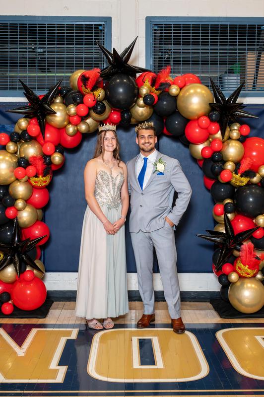 girl in fancy dress with crown and boy in suit with crown under balloon arch