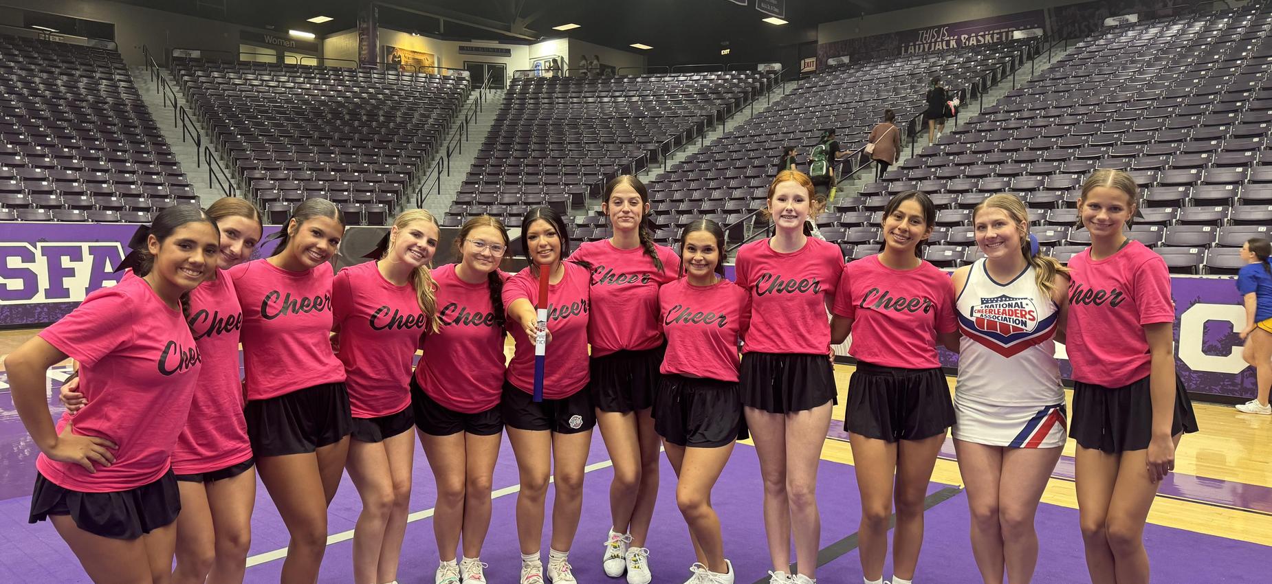 A group of cheerleaders wearing pink shirts poses together on a gym floor.