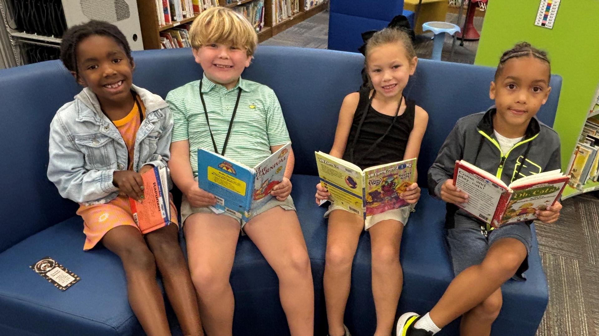 Four children sitting on a blue couch, happily reading books in a library setting.