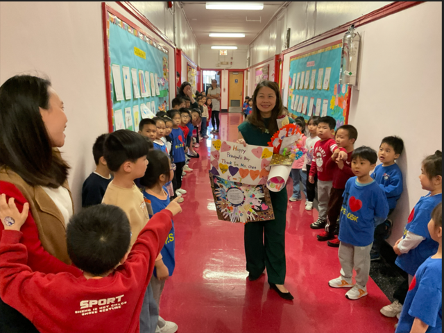 A woman stands in a colorful hallway holding flowers and a poster, surrounded by children.