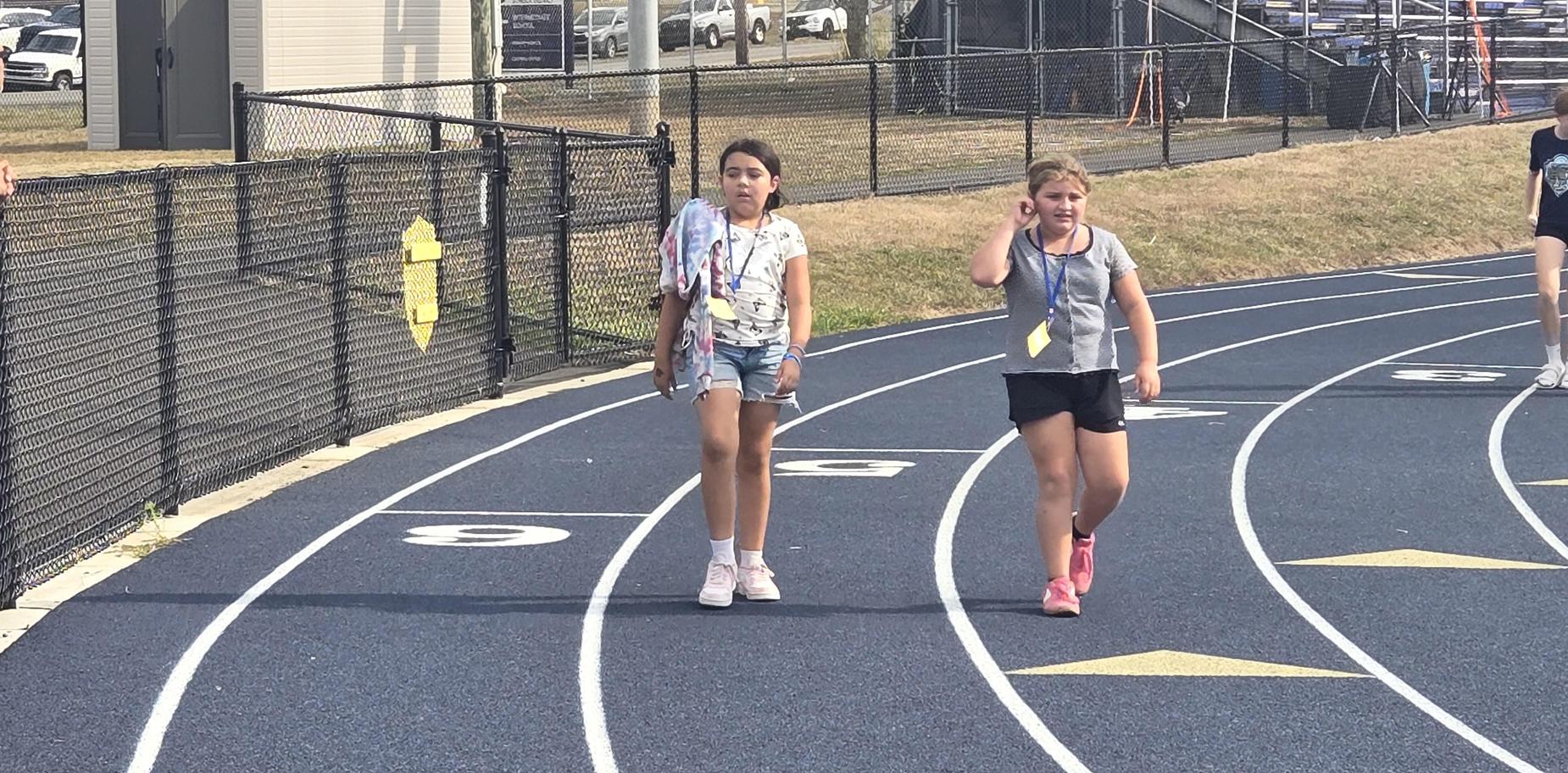 Two girls walking on the track with others running nearby, sunny day at a sports field.