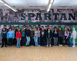 A group of high school students pose together in a school hallway beneath a large “SPARTAN” mural.