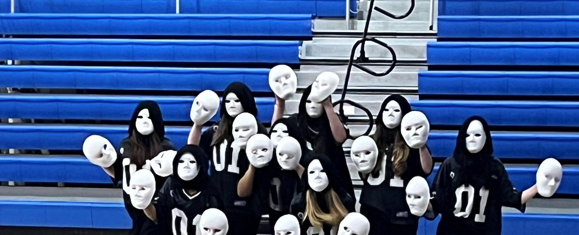 Group of dancers in black outfits holding white masks in a gymnasium.