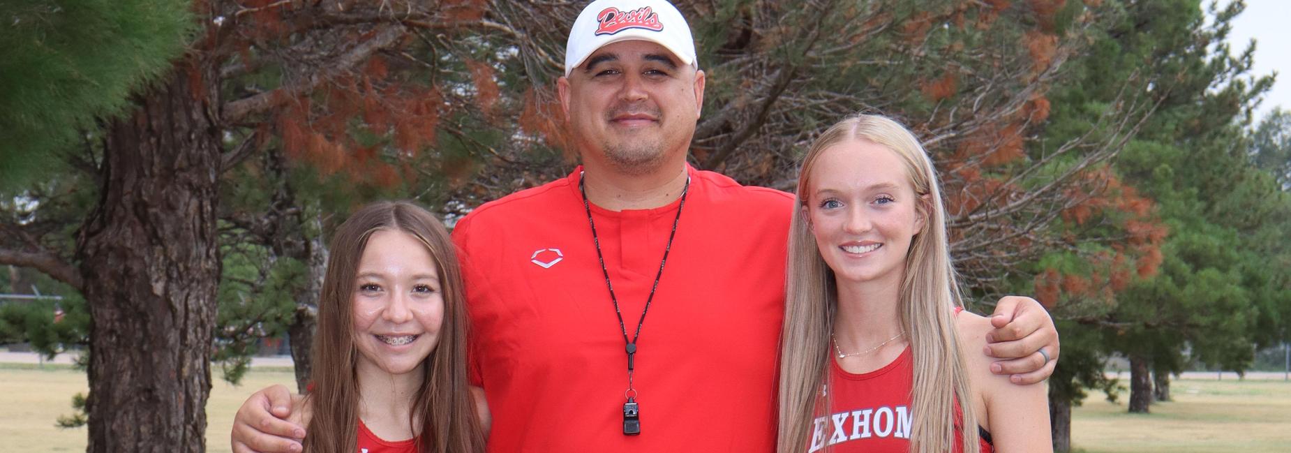 Two female athletes standing next to a coach, all in red uniforms on a grassy field.